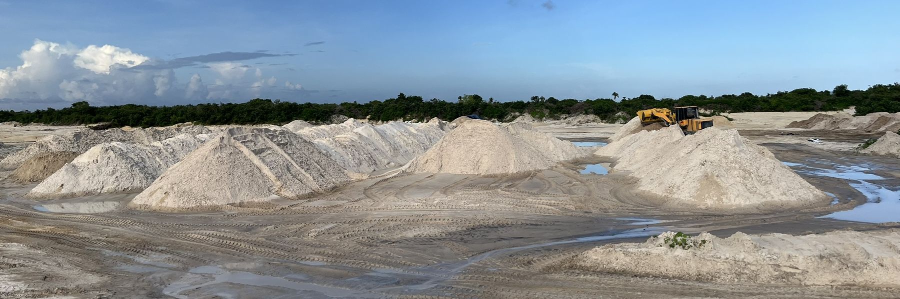 Pátio de estoque com montes de areia lavada e pá carregadeira, sob céu azul